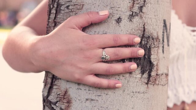 A view of a woman's hand with her ring and well-groomed nails, hugging a tree in a park on a sunny day, caring for and protecting it.