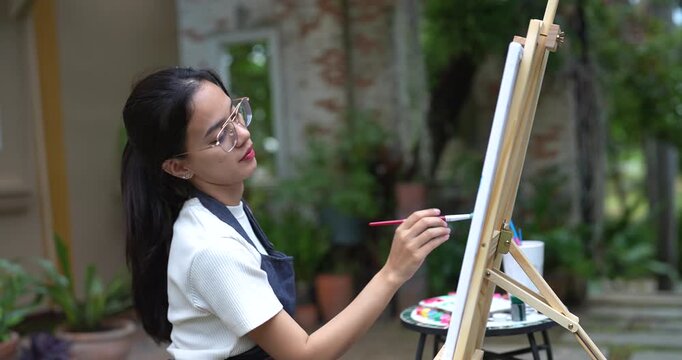 asian woman drawing ocean on canvas outdoors in tranquil backyard holding paintbrush with confident grip focused on creative process seated near greenery enjoying artistic hobby in natural quiet space