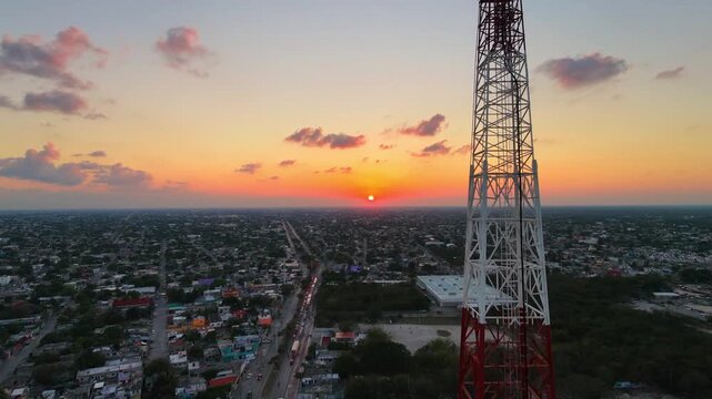 Cinematic 4K aerial ascent of a red and white cell tower against a vibrant orange sunset over Cancun, Mexico, showing city infrastructure and evening traffic.