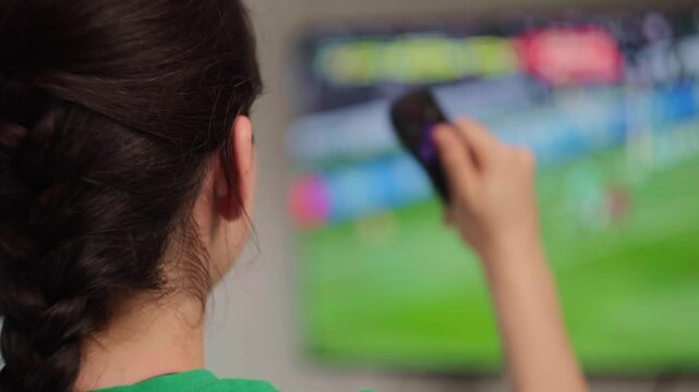 Woman with a braided hairstyle watching a soccer match on television and using a remote control to change channels or adjust the volume in the living room at home
