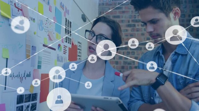 Woman tapping tablet, man leaning, causing network nodes linking tablet and wall charts for design