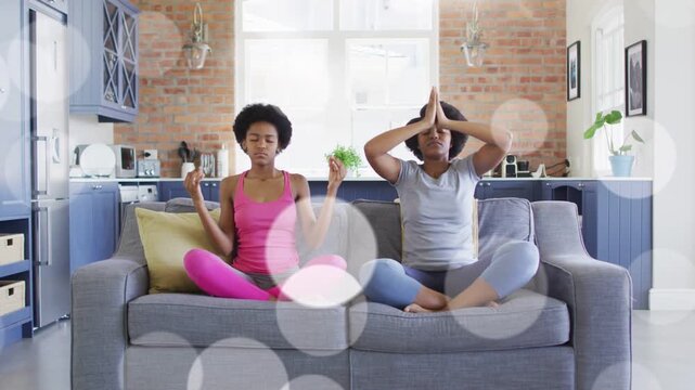Mom and daughter inhaling raising arms forming mudra on sofa for yoga with bokeh overhead