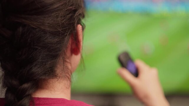 Woman with braided hair using a remote control to change channels while watching a soccer cup on a big screen television, enjoying her free time in a modern living room