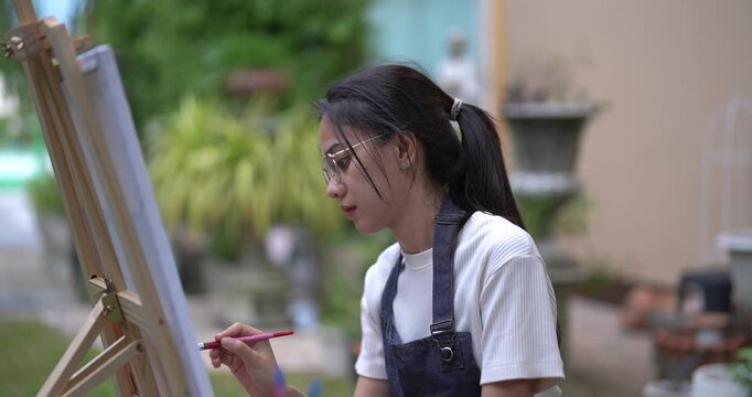asian woman sitting outdoor painting on canvas with relaxed focus holding brush during creative garden session enjoying tranquil hobby moment surrounded by plants and nature in peaceful backyard space
