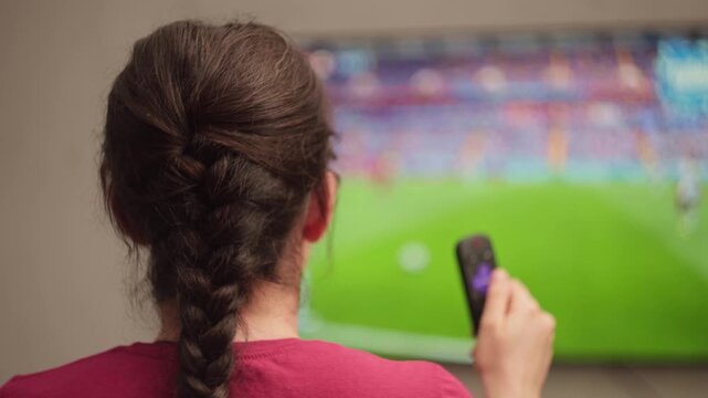 Woman with braided hair watching soccer match on television at home, holding a remote control and changing channels during a live broadcast in the living room