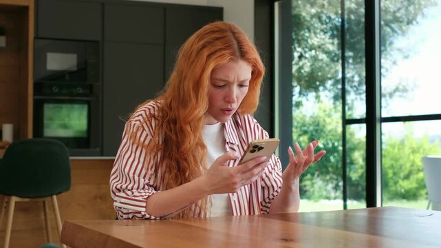 A young woman sitting at a table at home reacts with frustration and concern to something she sees on her smartphone. She appears upset by the news or message she is reading.