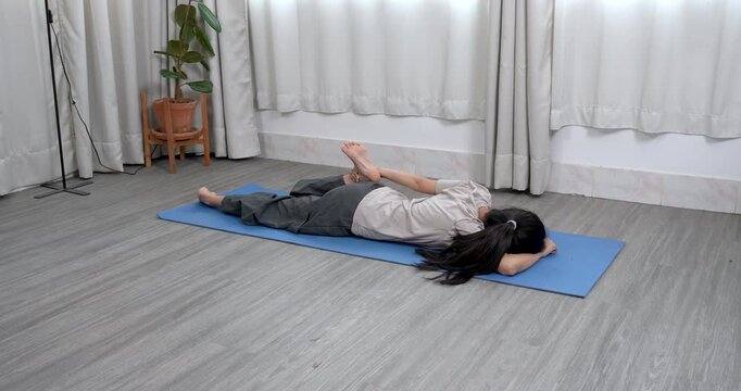 asian woman lying face down on yoga mat gently holding one foot behind during quad stretch in relaxing home routine to improve flexibility reduce tension and enhance body awareness