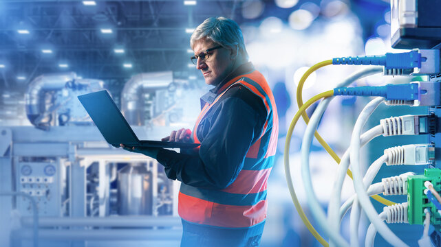 Industrial automation, Equipment setup. Engineer configures production systems with laptop, adjusting machinery and network connections in modern factory.