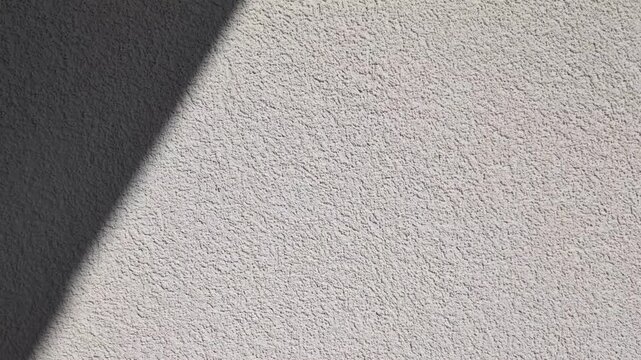 Cinematic pan of wall texture with sunlight shadow, minimalist plaster surface, vertical camera movement, candid style video, architectural detail, grainy cement backdrop, abstract light and shadow.