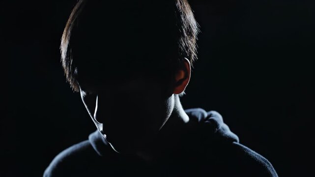 Dramatic rim lighting outlines the silhouette of a person in a hooded sweatshirt against a solid black background, face hidden in deep shadow, cinematic low key lighting style.