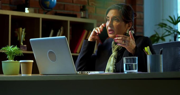 Senior Indian businesswoman in corporate attire attending important phone call on smartphone while sitting at workstation with laptop in cozy office space experienced female executive at desk