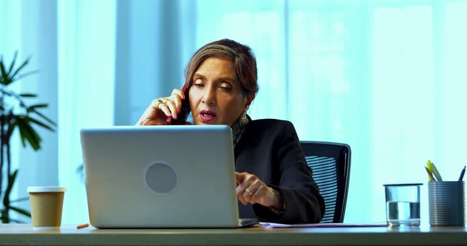 Senior Indian businesswoman in corporate attire attending important phone call on smartphone while sitting at workstation with laptop in cozy office space experienced female executive at desk