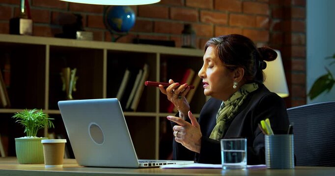Senior Indian businesswoman in corporate attire attending important phone call on speaker phone via smartphone at workstation with laptop in cozy office space experienced female executive desk