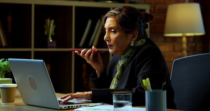 Senior Indian businesswoman in corporate attire attending important phone call on speaker phone and typing on laptop in modern cozy office space experienced female executive with white hair