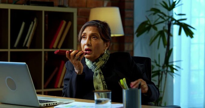 Senior Indian businesswoman in corporate attire attending important phone call on speaker phone and typing on laptop in modern cozy office space experienced female executive with white hair
