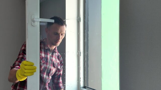 Caucasian man in yellow rubber gloves diligently cleaning a large window pane with a professional squeegee during household chores