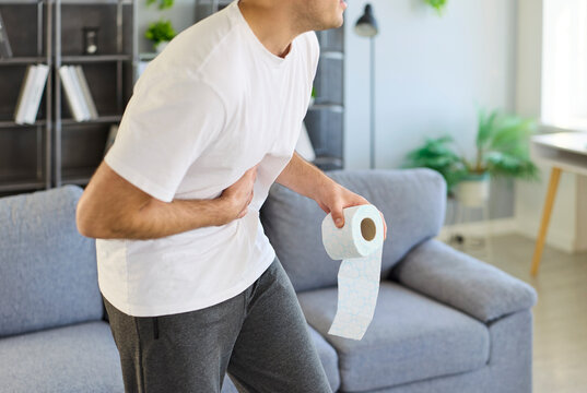 Man with diarrhea clutching abdomen in pain. He bends with stomachache and cramps, holding toilet paper. It shows urgency, nausea, and abdominal discomfort. Clear concept of urgent digestion.
