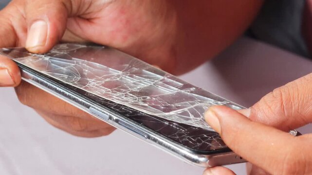 Skilled repairman removing shattered tempered glass screen protector from cellphone, revealing intact phone screen underneath.