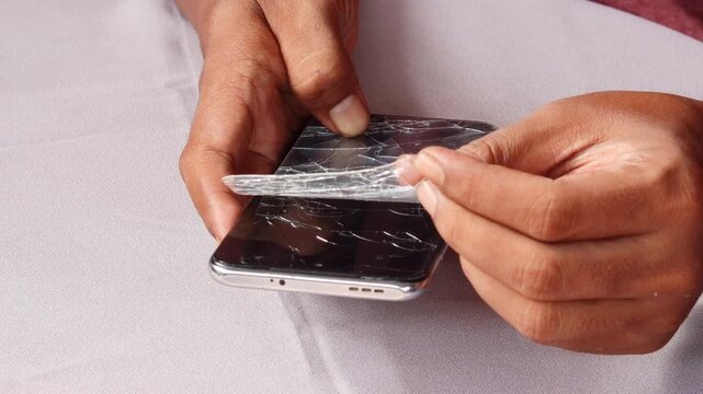 Skilled repairman removing shattered tempered glass screen protector from cellphone, revealing intact phone screen underneath.