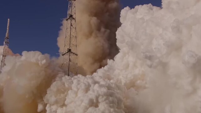A massive rocket engine ignites at the launchpad creating thick clouds of smoke and fire during a daytime liftoff.