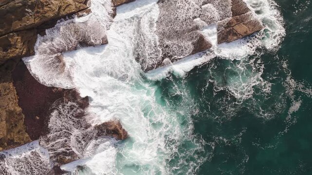 Stunning top down drone footage remains static over the rugged coastline as powerful white ocean waves crash and churn against ancient sandstone rocks creating beautiful sea foam patterns