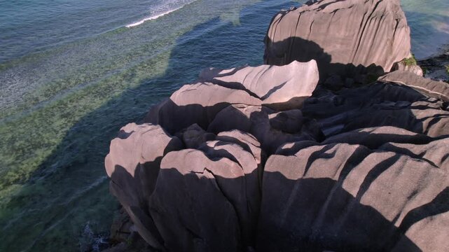 Aerial shot gliding over sculpted granite boulders to reveal the crystal clear turquoise waters and coral reefs of Anse Grosse Roche on La Digue Island, Seychelles. 013