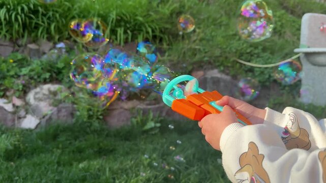 Close-up of a child using a colorful toy to create shimmering soap bubbles outdoors in a sunny garden, capturing playful childhood moments, leisure, and family fun in nature.