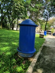 Obraz premium Blue cylindrical trash bin in a park with lush green grass and trees container cylinder municipal cleanliness daylight