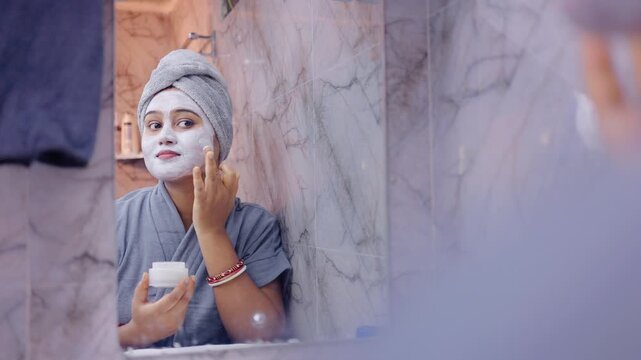 Close up shot of a woman applying face pack while holding a cosmetic jar in front of a bathroom mirror.