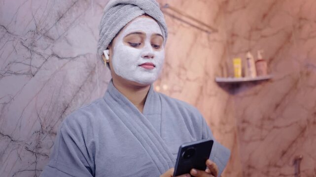 Female applying face mask and using earbuds to listen music on smartphone, standing in the modern bathroom.