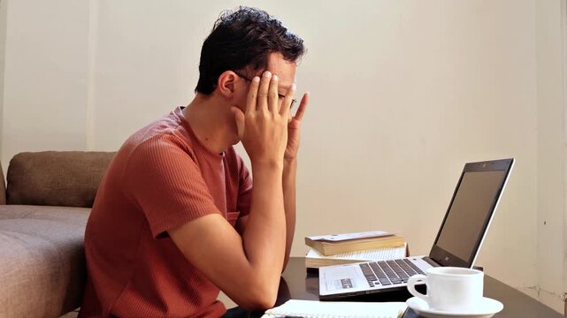 An Asian man holding his head while working on a laptop at home, showing stress, headache, and mental pressure. This scene represents work overload, burnout, frustration, and challenges in remote work