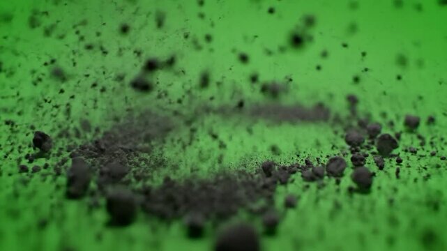 A small pile of dark, granular material explodes on a bright green surface, creating a dynamic visual effect and abstract composition