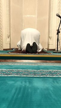 Static vertical shot of an Imam in white traditional clothing sitting in prayer within a beautifully carved stone Mihrab. Spiritual and serene atmosphere.