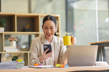 Young Asian businesswoman smiling while checking social media on smartphone, holding a yellow mug,...