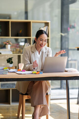 Professional Asian woman smiling, engaged in a virtual meeting on her laptop at an office desk,...