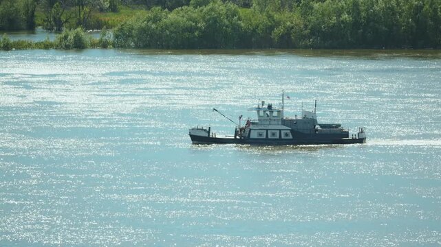 A small industrial workboat moves across a sunlit river. Bright sun creates sparkling reflections on the water. The distant green shore provides a natural background. Calm transportation scene