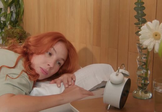 Sleepy woman reaching for alarm clock in bed, resisting early morning wakeup