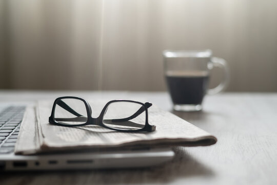 Reading glasses resting on a folded newspaper over a laptop, with a glass mug of black coffee in the soft background, depicting a morning routine of information consumption