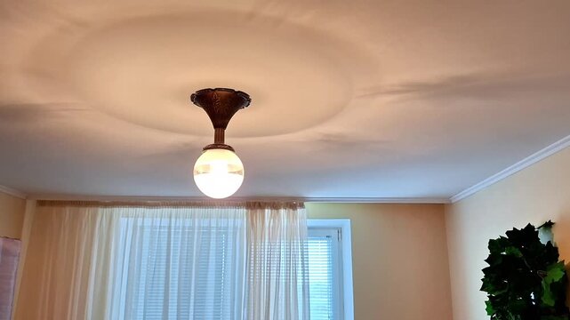 Round white bulb ceiling light with decorative brown base glows softly above sheer curtains and green plant, creating a warm, cozy indoor atmosphere with natural daylight filtering in