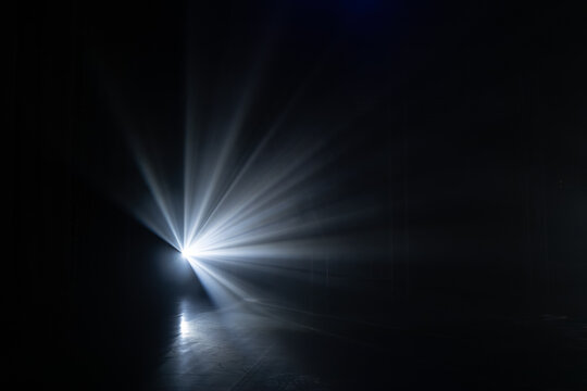 Bright light beam projecting from a dark background, creating dramatic rays and shadows on a smooth floor surface in a dimly lit environment