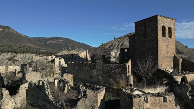 Desolate ruins of the abandoned medieval village of Esco in Aragon, Spain, featuring the remnants of old stone houses and a historic church tower against a backdrop of barren hills