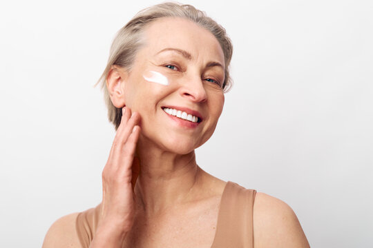 Smiling mature woman with short grey hair applies white face cream on cheek. Natural skincare concept with healthy glowing skin and anti aging beauty routine on white studio background.