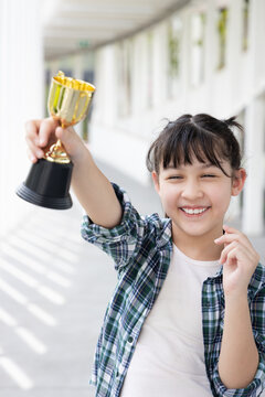 Confident, happy smiling young Asian grade-schooler or primary school girl raising a shiny gold trophy cup up high, concept image for learning success, academic achievement