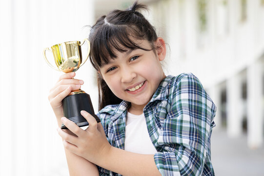 Confident, happy smiling young Asian grade-schooler or primary school girl holding a shiny gold trophy cup,  concept image for learning success, academic achievement, contest winning prize