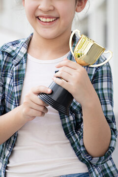Hand closeup of confident, happy smiling young Asian grade-schooler or primary school girl holding a shiny gold trophy cup, concept image for learning success, academic achievement