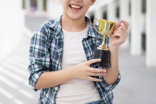 Hand closeup of confident, happy smiling young Asian grade-schooler or primary school girl holding a shiny gold trophy cup, concept image for learning success, academic achievement