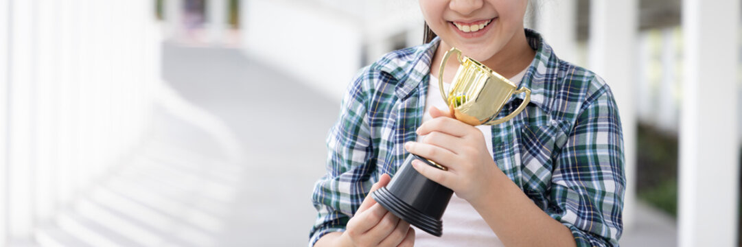 Hand closeup of confident, happy smiling young Asian grade-schooler or primary school girl holding a shiny gold trophy cup, concept image for learning success, academic achievement