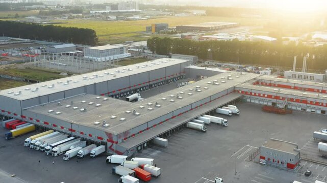 Aerial Drone View: Large Modern Logistics and Distribution Hub with Trucks at Loading Docks at Golden Hour Sunset | Warehouse Complex and Freight Transportation Concept