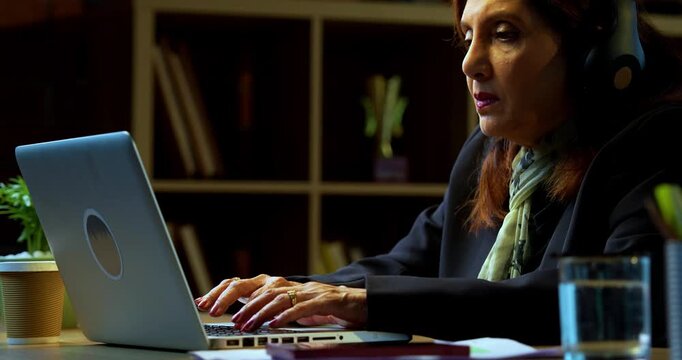 Senior Indian businesswoman in corporate attire typing on laptop with headphones on at workstation in modern cozy office space experienced female executive with white hair working at her desk