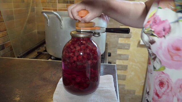 Close-up of female hands closing a glass jar with homemade cherry compote, Traditional process of home canning, sealing a large glass jar with red cherry fruit compote for winter storage.